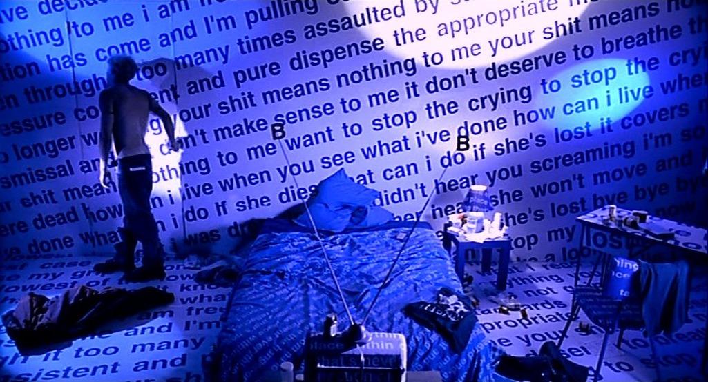 Black text is scrawled endlessly along a white wall. The room is lit blue with a blue bed in the middle, and a small tv with a large antenna in front. On the left a man stands, topless reaching for the door, his back to us.