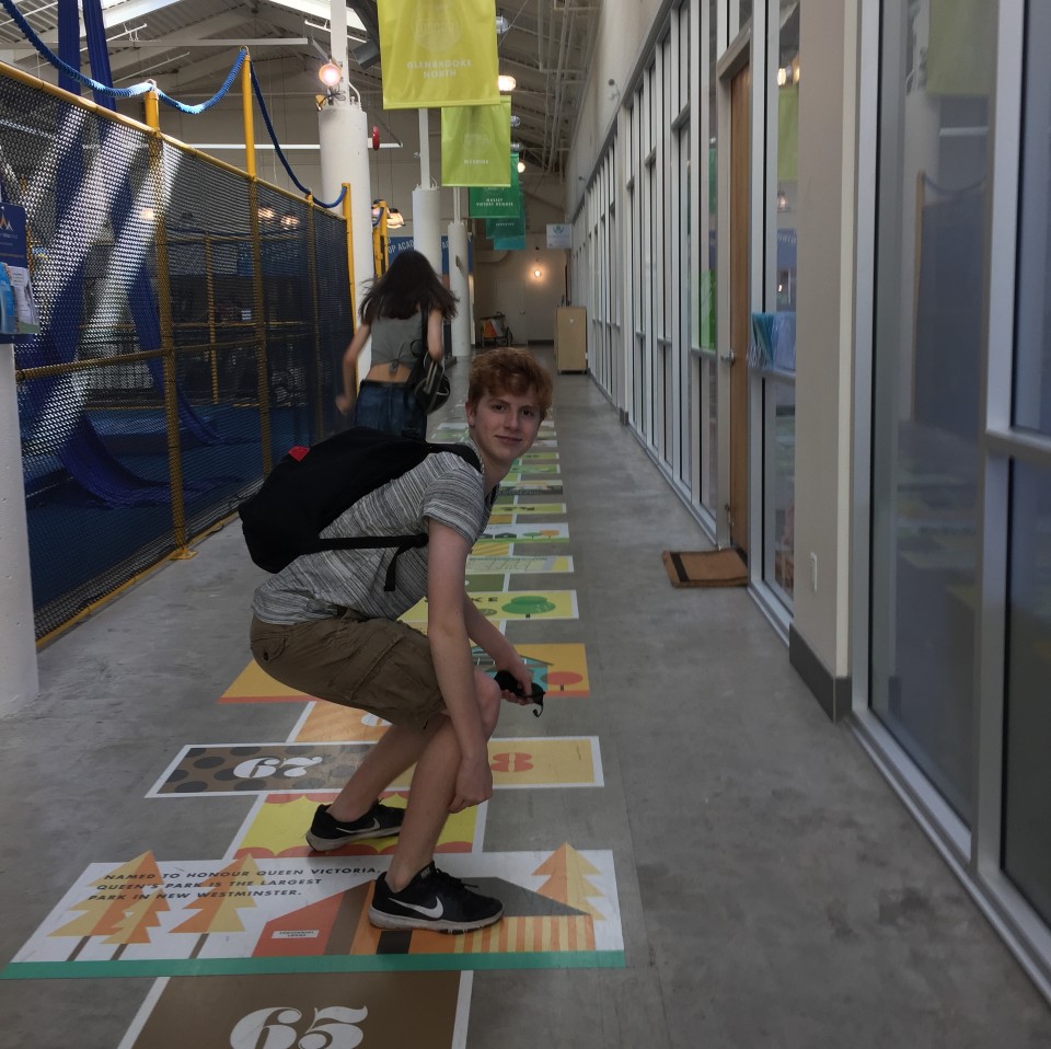a young red-haired boy stands crouched in a long hallway facing us. He is wearing a grey striped shirt, khaki brown cargo shorts, black nike running shoes, and a black backpack. On the floor of the hallway is a hopscotch grid with fun facts. Behind him is a brunette woman in a grey tshirt and jeans running away while holding a large black purse. On the left side there is a blue and yellow play structure, and on the right there are rows of windows, spanning down the hall.