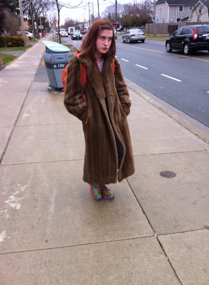 A reddish-brown haired teenage girl stands next to the curb of a busy street in an orange backpack, lavender shirt, colourful shoes, and a large fur coat. She looks right, and behind her is a city trash bin.
