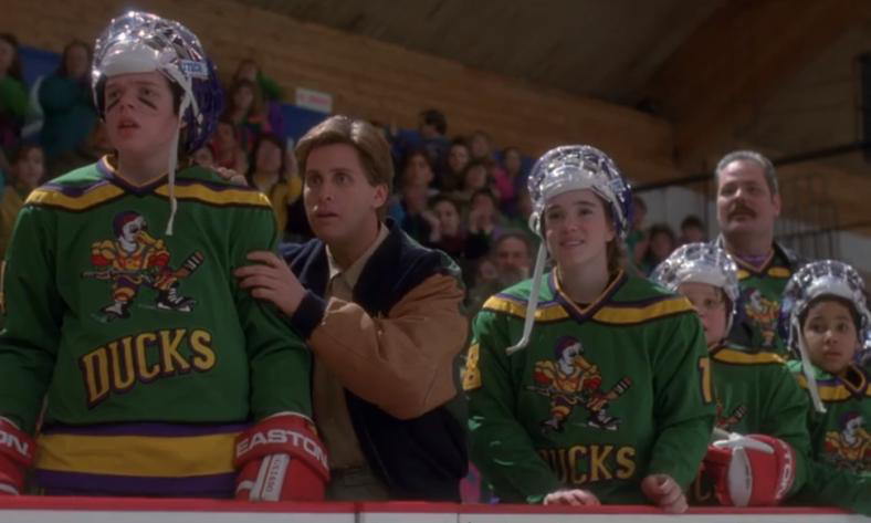 Movie still from The Mighty Ducks. Teen hockey players watch a game from the sidelines.