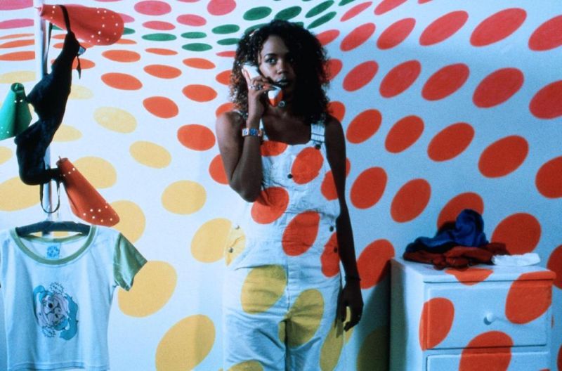 Against a background with multicoloured twister-esque spots, a dark-skinned young woman dressed all in white stands on the phone.