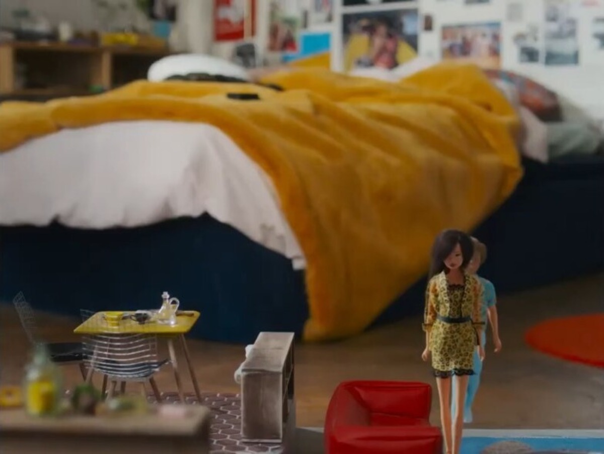 Inside a bedroom, a large bed with a blue base and yellow blanket. In the foreground is a miniature furniture set up for two dolls, one brunette in a cheetah print suit, and another, obscured by the first.