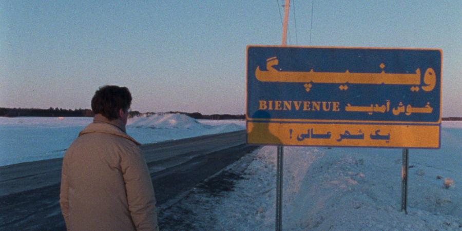 Movie still from Universal Language. A man stands looking at a snowy road, with a sign welcoming him in different languages.