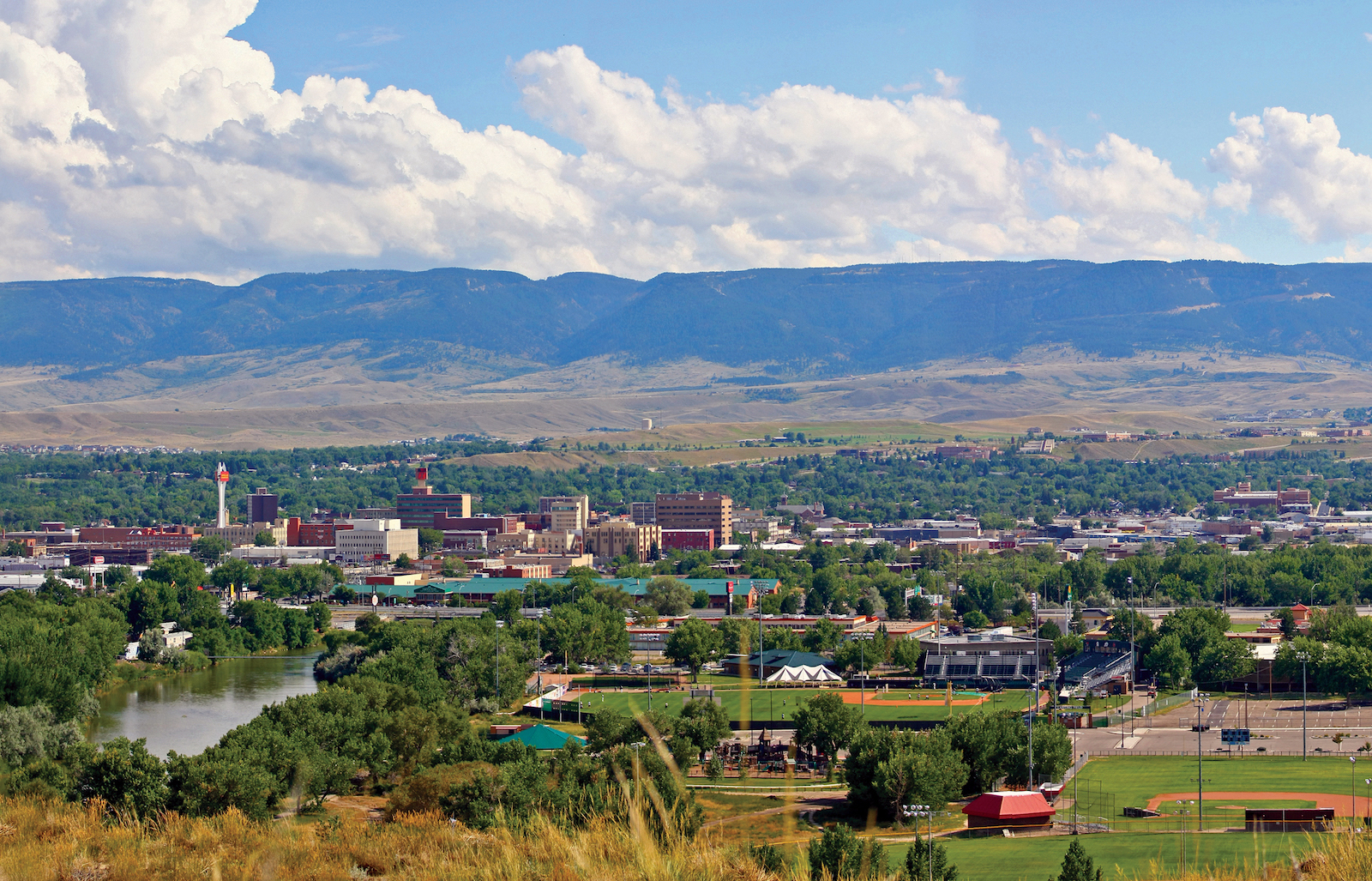 Aerial view of Casper, Wyoming city with river, trees, sports fields, and mountains under cloudy sky.