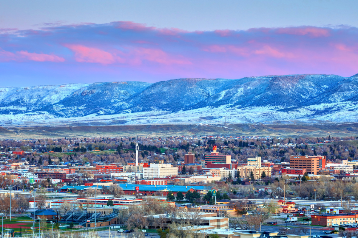 Cityscape with buildings and trees in the foreground and snow-covered mountains under a pink and blue sky at sunset.