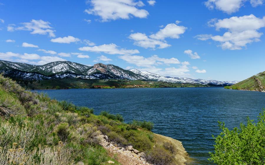 View of a large blue lake surrounded by green hills and snow-capped mountains under a partly cloudy blue sky.