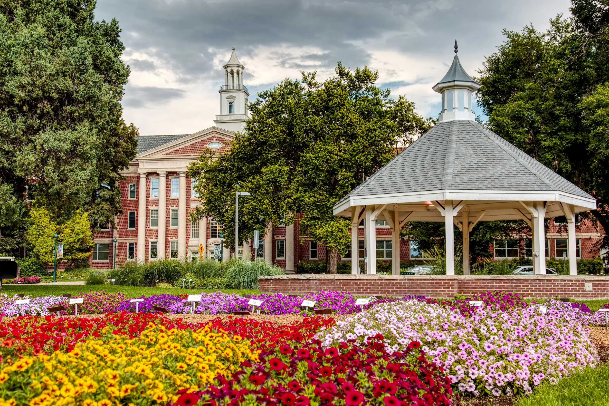 Colorful flower gardens in front of a brick building with white columns and two cupolas, alongside a white gazebo under a cloudy sky.