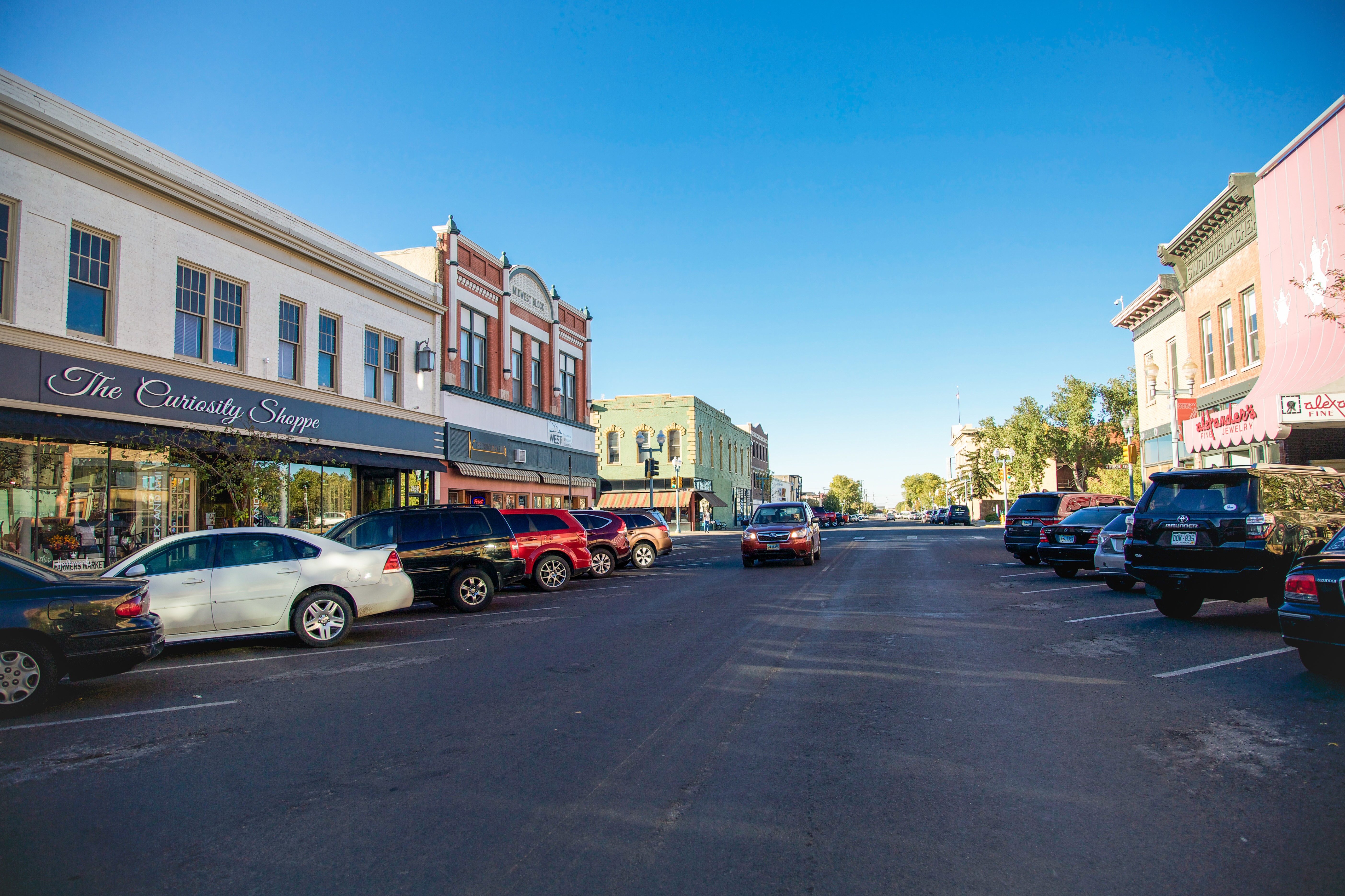 Downtown street scene with parked cars along both sides and buildings including The Curiosity Shoppe under blue sky.