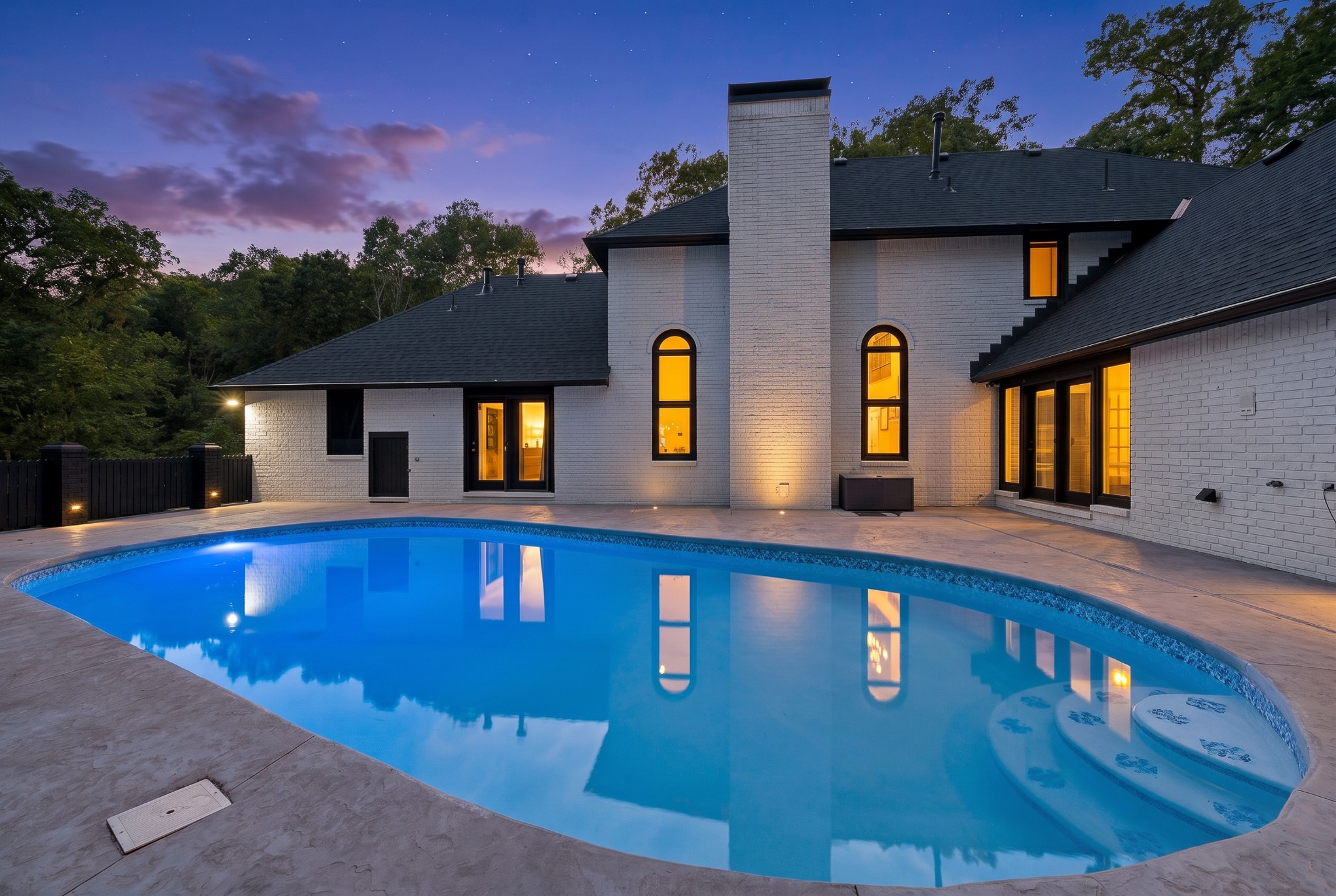 White brick home with illuminated windows and a blue swimming pool at twilight.