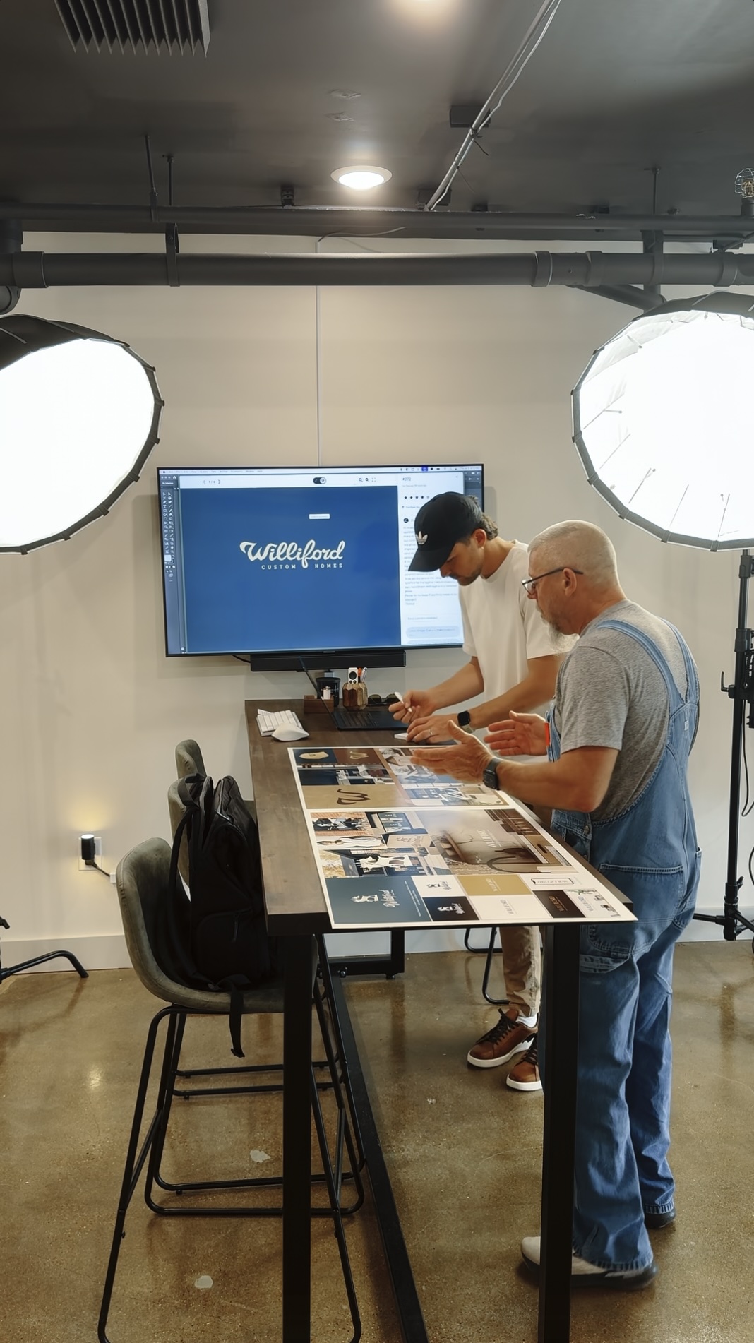 Two men reviewing printed design layouts on a tall table in a modern studio lit by two softbox lights.