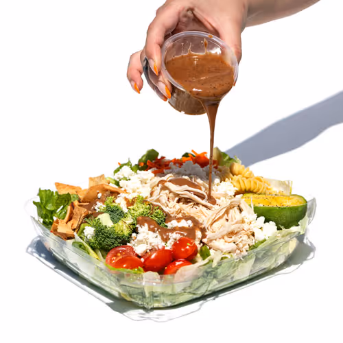 Hand pouring brown dressing over a fresh salad with chicken, cherry tomatoes, broccoli, pasta, avocado, and feta cheese in a clear plastic container.