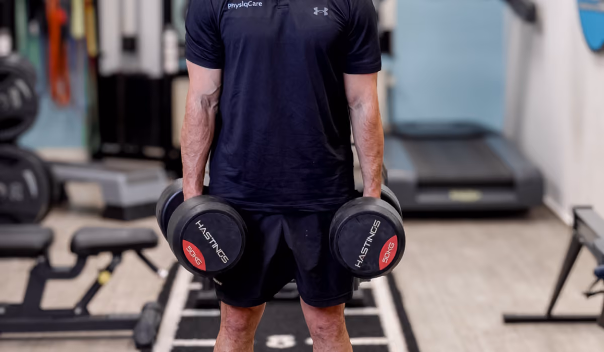 Man in PhysiqCare-sportkleding staat in de fitnessruimte met twee zware dumbbells in zijn handen voor krachttraining.