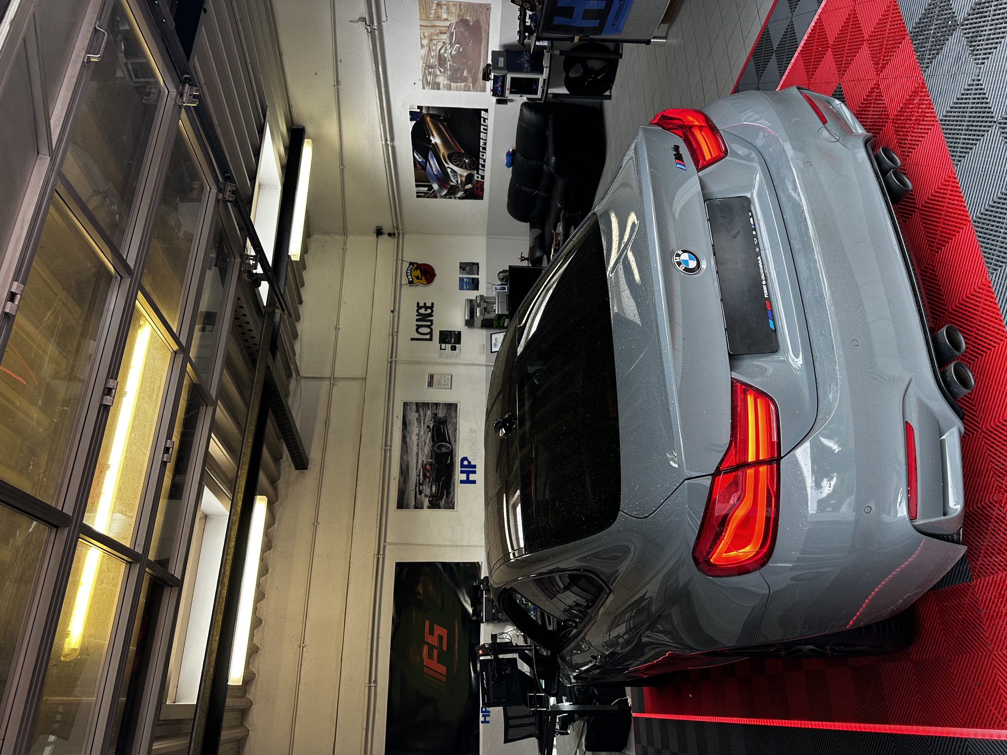 Gray BMW M4 parked on red and black checkered floor inside a garage with posters on walls.