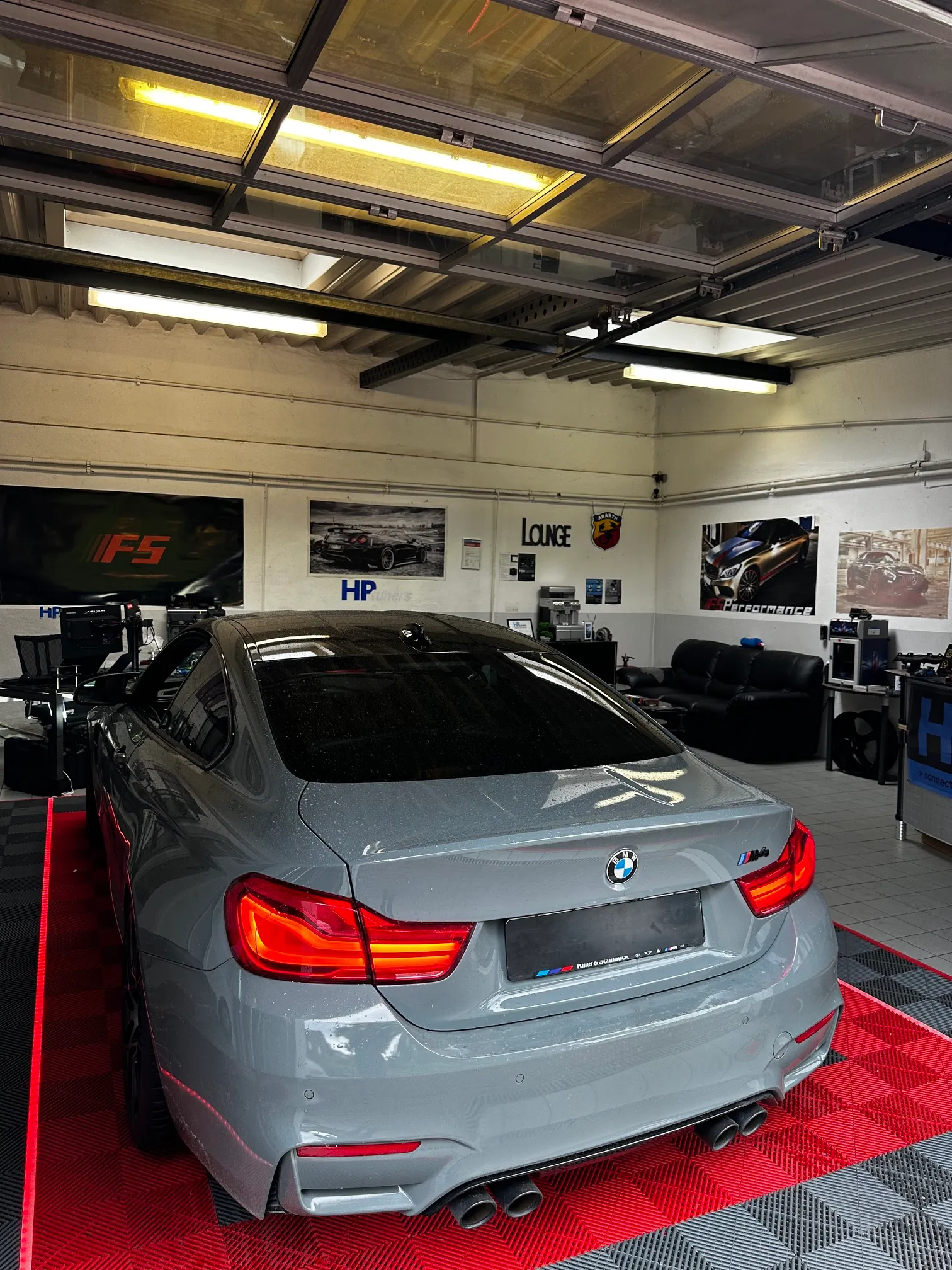 Gray BMW M4 parked on red and black checkered floor inside a garage with posters on walls.
