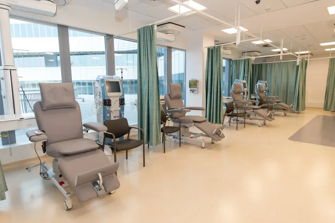 Modern dialysis unit with multiple reclining treatment chairs separated by green privacy curtains and dialysis machines.