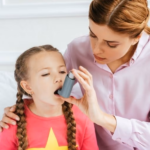 Woman helping a young girl use an asthma inhaler with her mouth open.