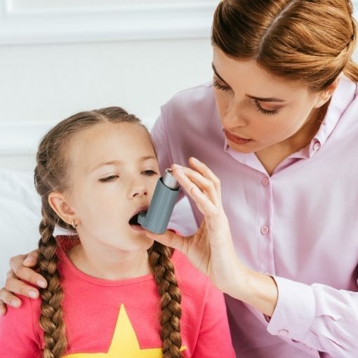 Woman helping a young girl use an asthma inhaler with her mouth open.