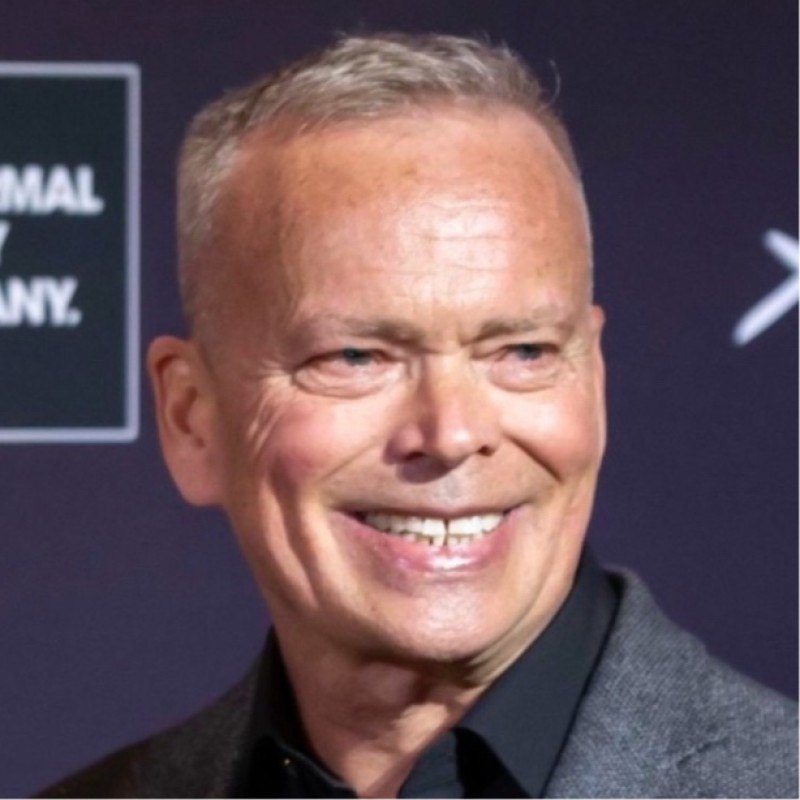 Smiling middle-aged man with short grey hair wearing a dark shirt and grey blazer against a dark background.