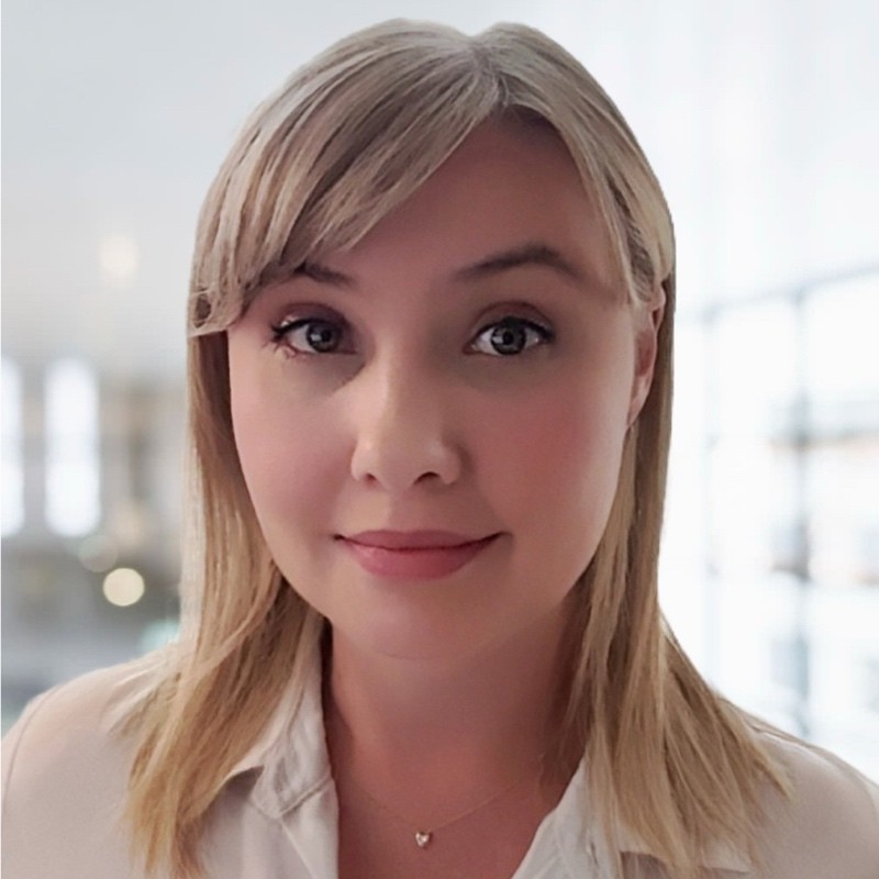 Portrait of a smiling woman with straight blonde hair wearing a white shirt and a heart-shaped necklace in a bright indoor setting.
