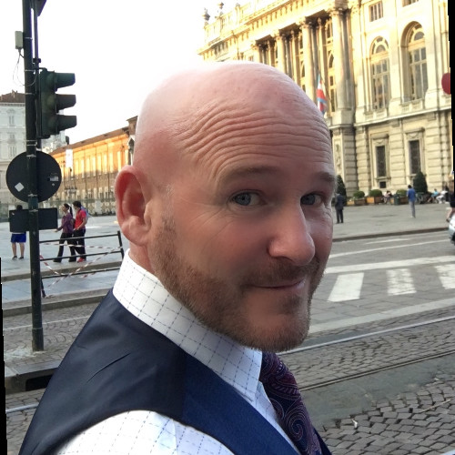 Bald man wearing a white shirt, blue vest, and patterned tie looking back while standing on a cobblestone city street.