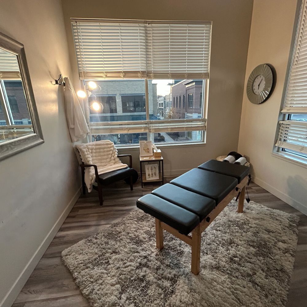 Calm therapy room with a black massage table, a cozy chair draped with a white blanket, a small side table, and large windows with blinds partially open.