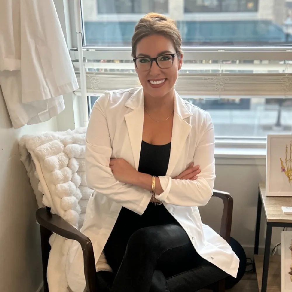 Dr. Jess, Chiropractic Specialist, wearing glasses and a white lab coat sitting cross-legged in a chair in a medical office.