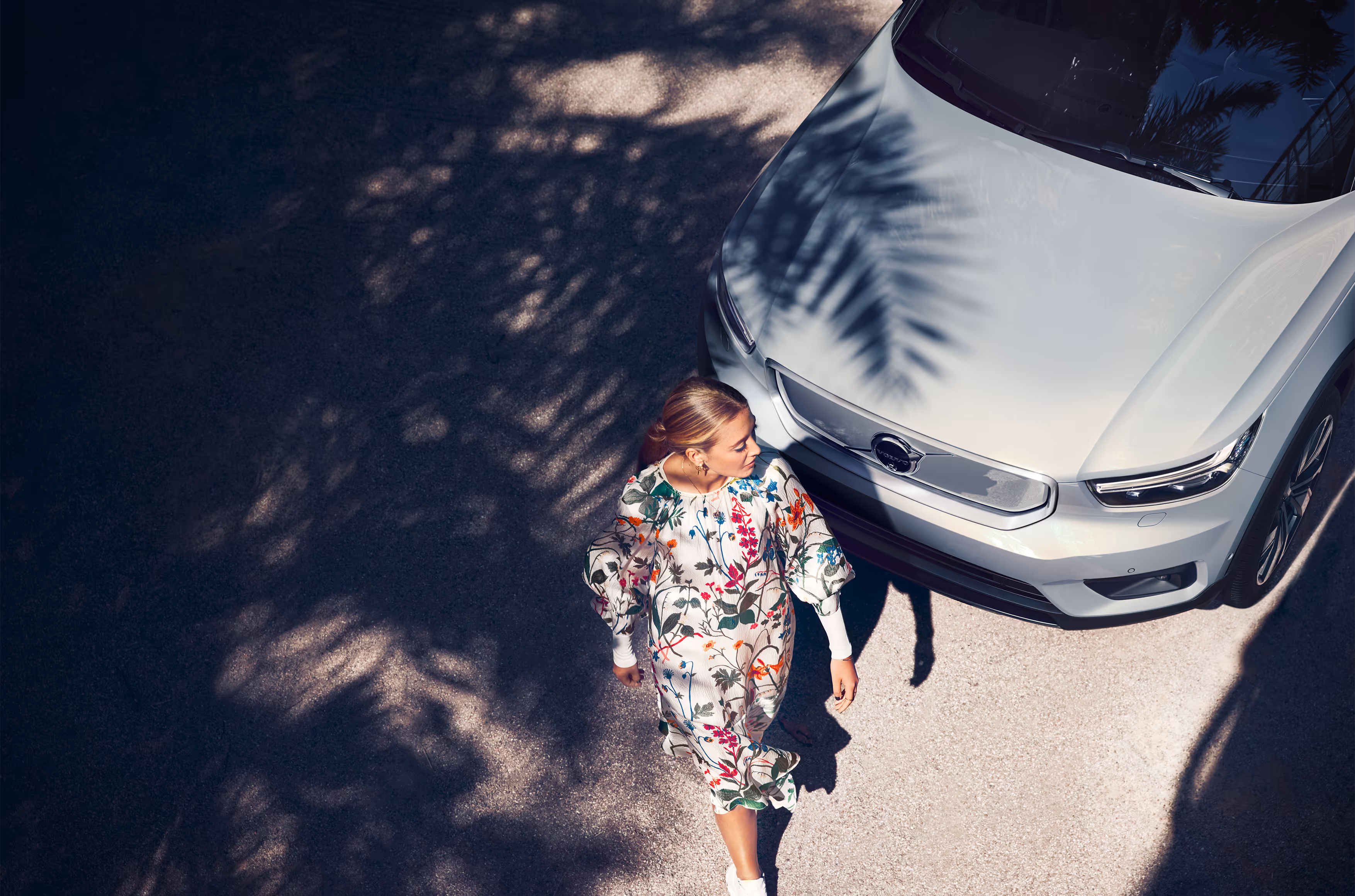 Woman in a floral dress walking beside a white Volvo car with palm tree shadows on the ground.