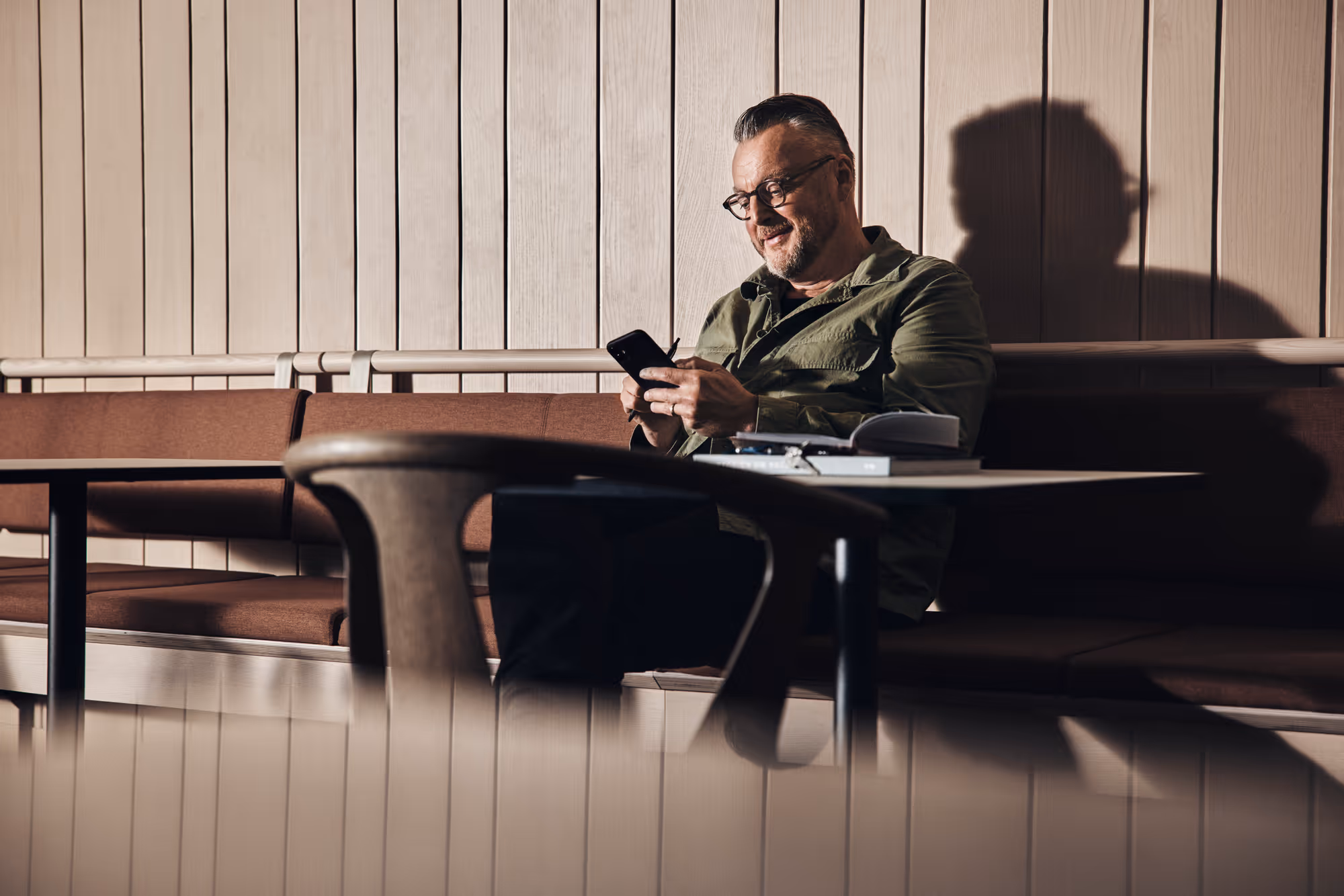 Man sitting on a brown cushioned bench using a smartphone, with a book and glasses on the table in front of him.