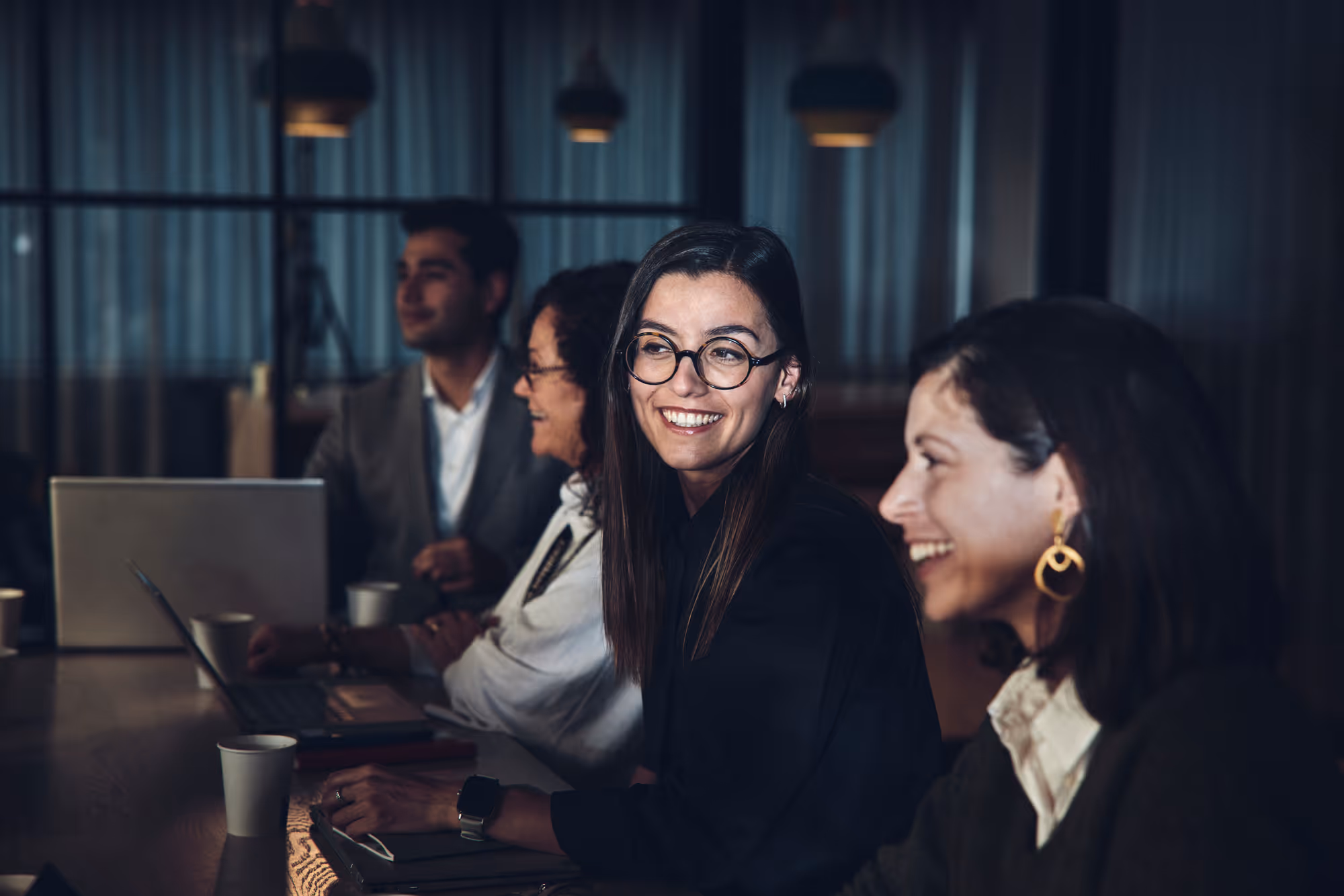 Four diverse professionals sitting at a table with laptops, smiling and engaging in a meeting in a dimly lit office.
