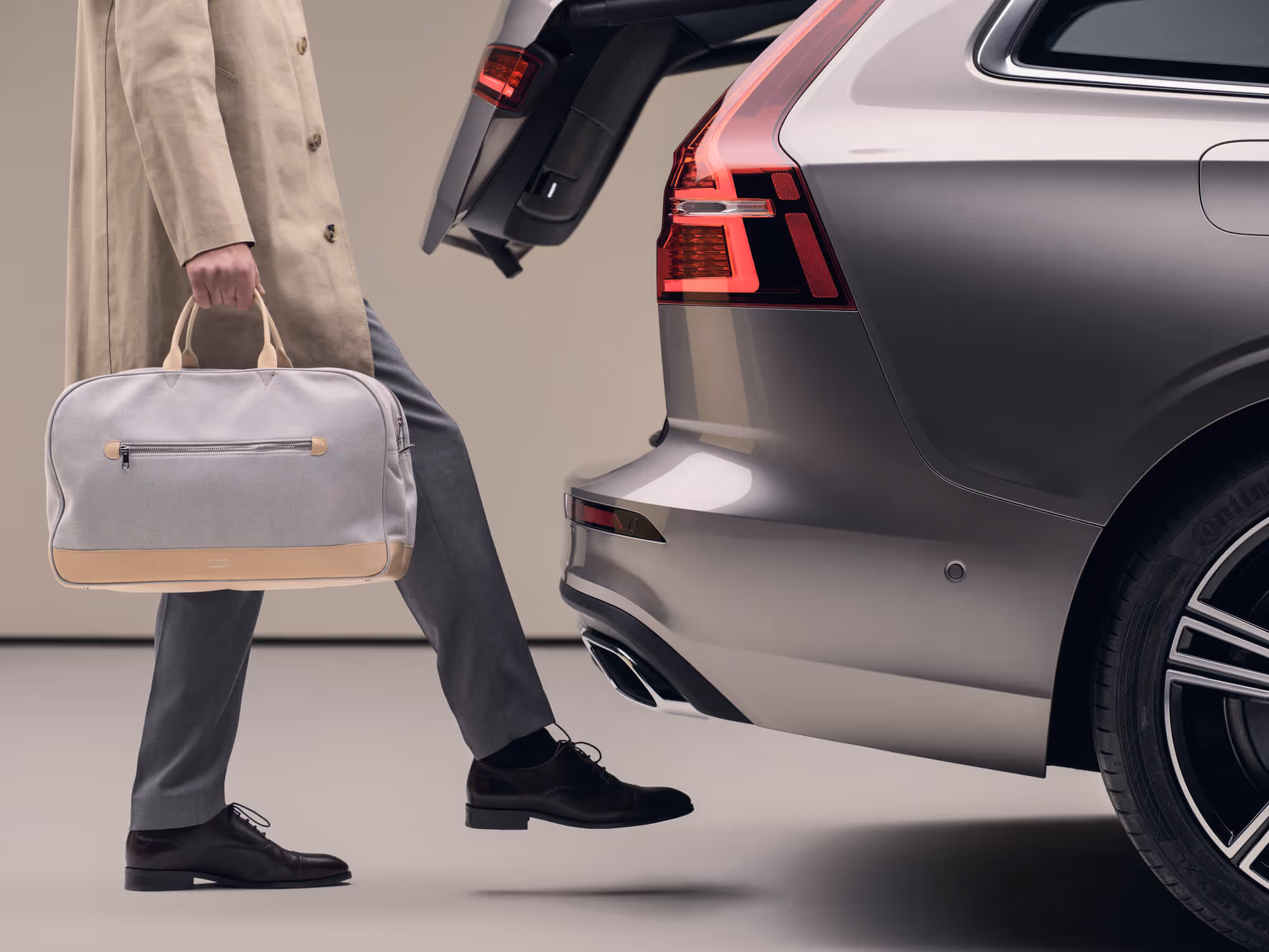 Person with beige trench coat carrying a gray duffel bag, stepping towards the open rear trunk of a gray car.