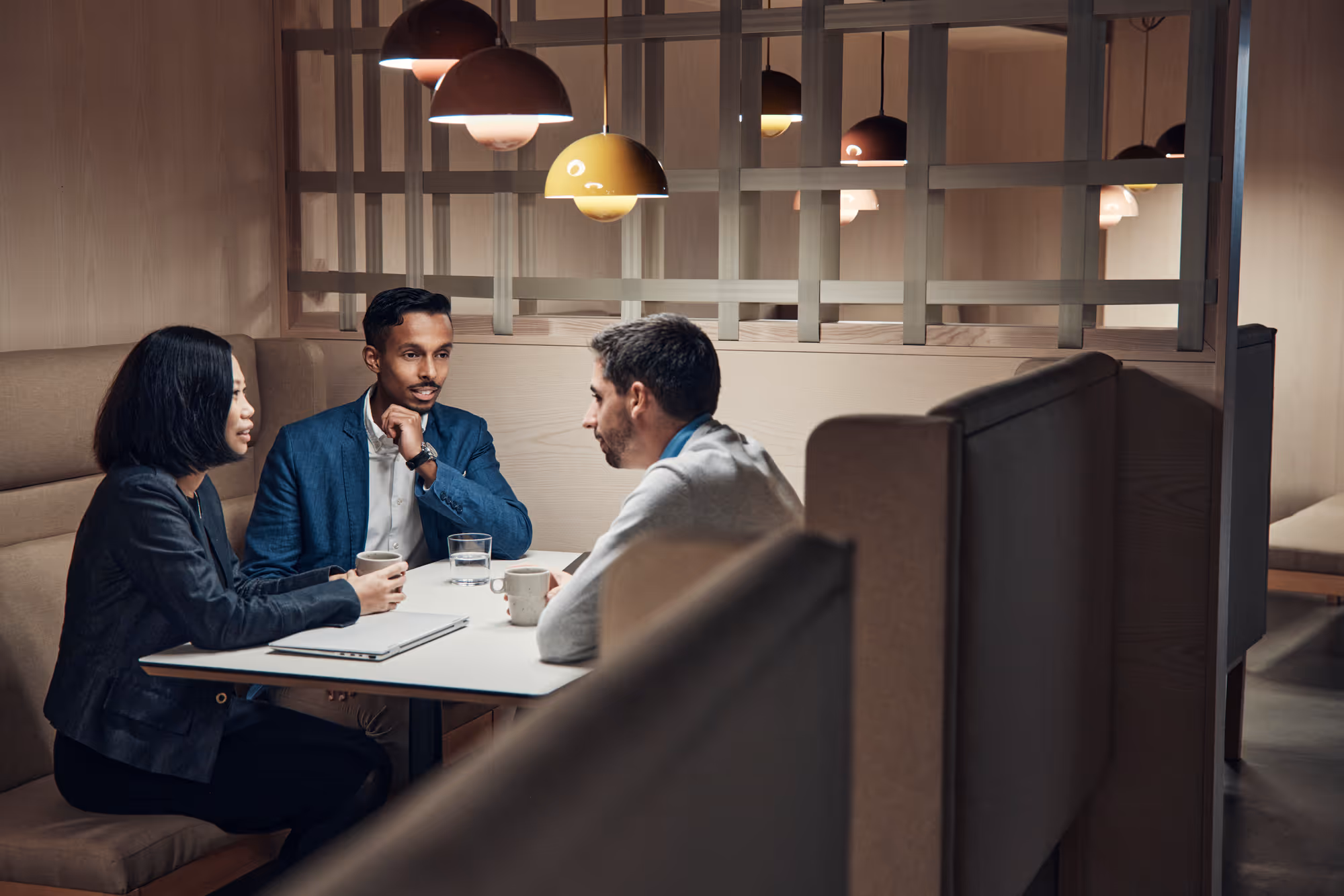 Three professionals having a discussion over coffee at a booth table in a modern office lounge with pendant lights.