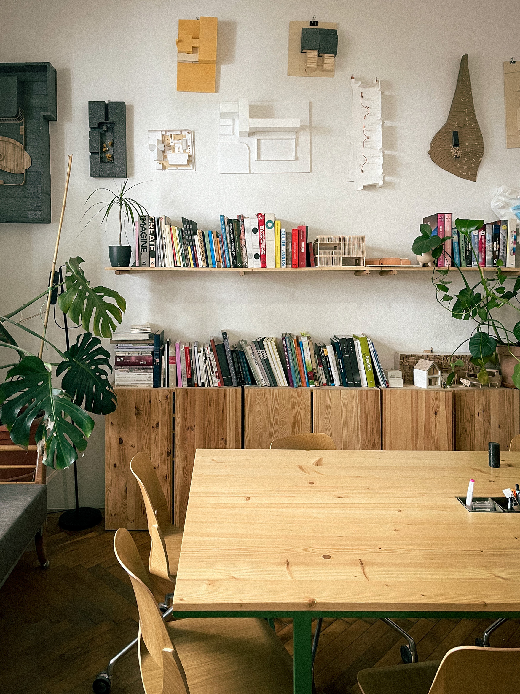 Cozy workspace with wooden table and chairs, bookshelves filled with books, architectural models on the wall, and large green plants.