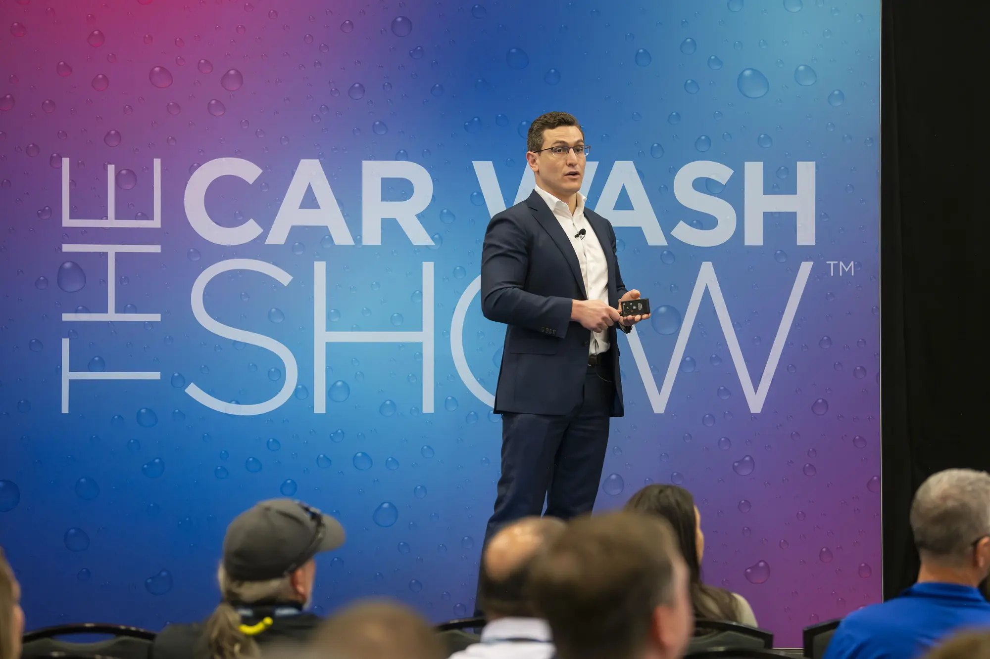 Man in suit speaking on stage at The Car Wash Show with audience in foreground.