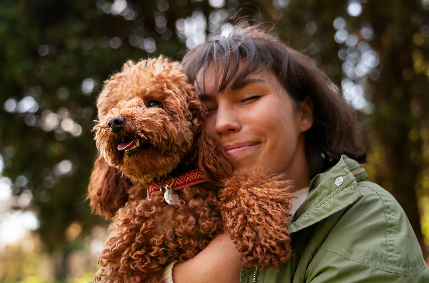 Woman in green jacket smiling with eyes closed hugging a happy curly brown dog outdoors.