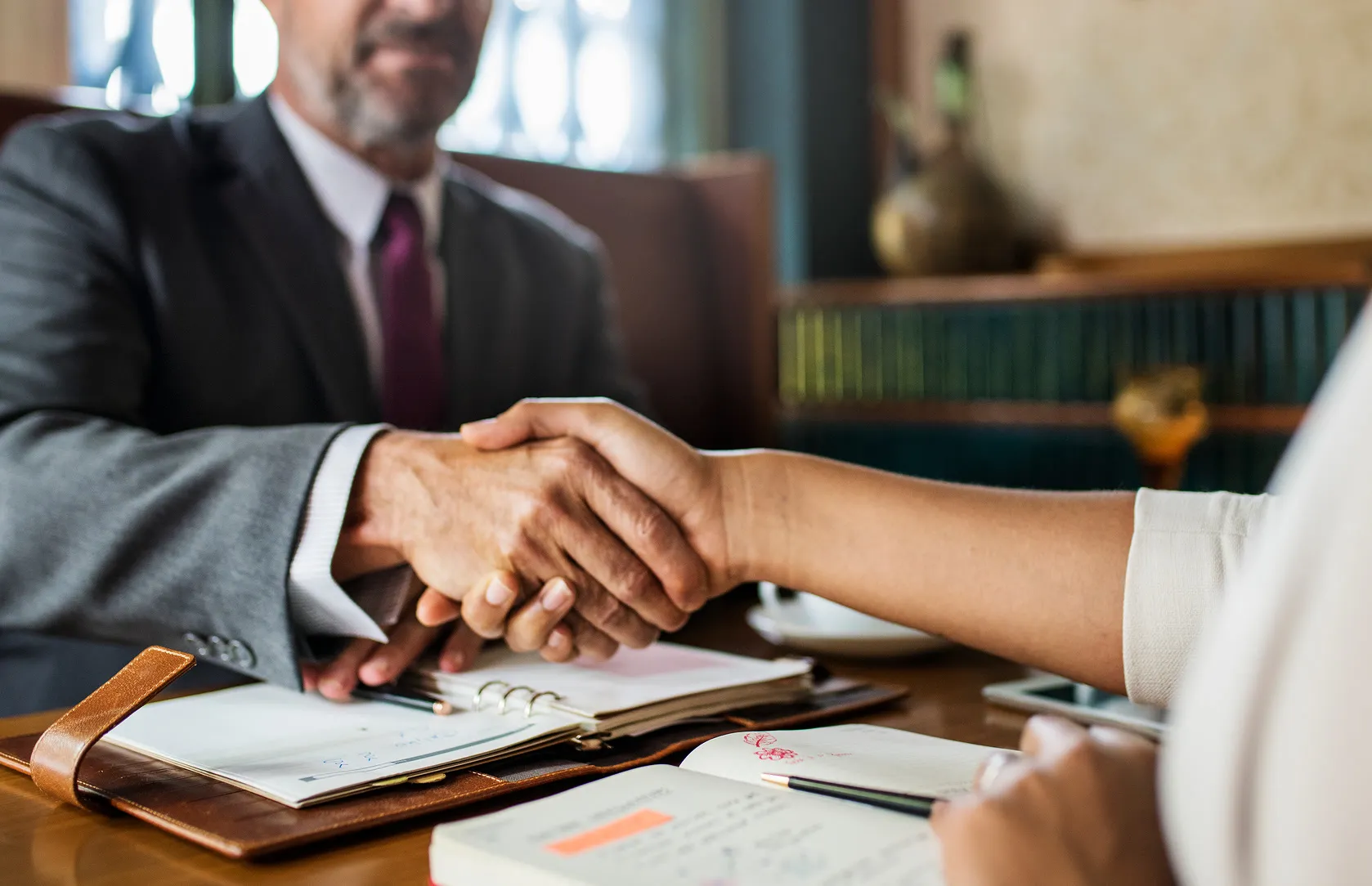 Two people shaking hands over a desk with open notebooks and a pencil during a meeting.