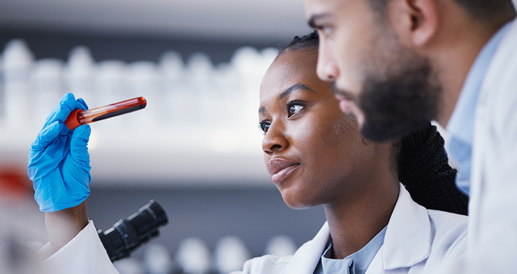 Two scientists in white lab coats examine a blood sample in a laboratory. In the foreground, a woman wearing blue nitrile gloves holds up a vial of blood, looking at it closely with a focused expression. A male colleague looks on from behind her. A microsc