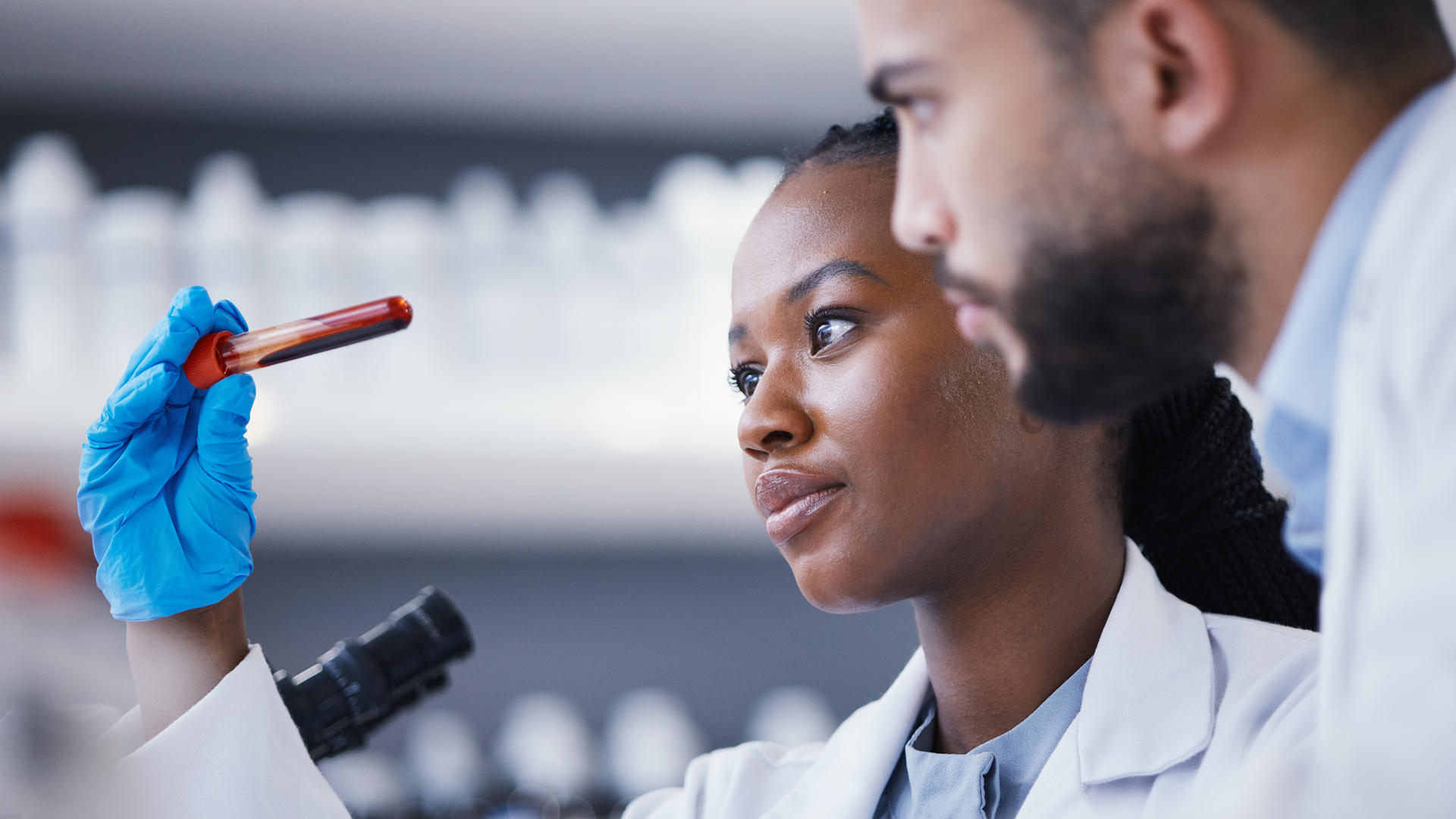 Two scientists in white lab coats examine a blood sample in a laboratory. In the foreground, a woman wearing blue nitrile gloves holds up a vial of blood, looking at it closely with a focused expression. A male colleague looks on from behind her. A microsc