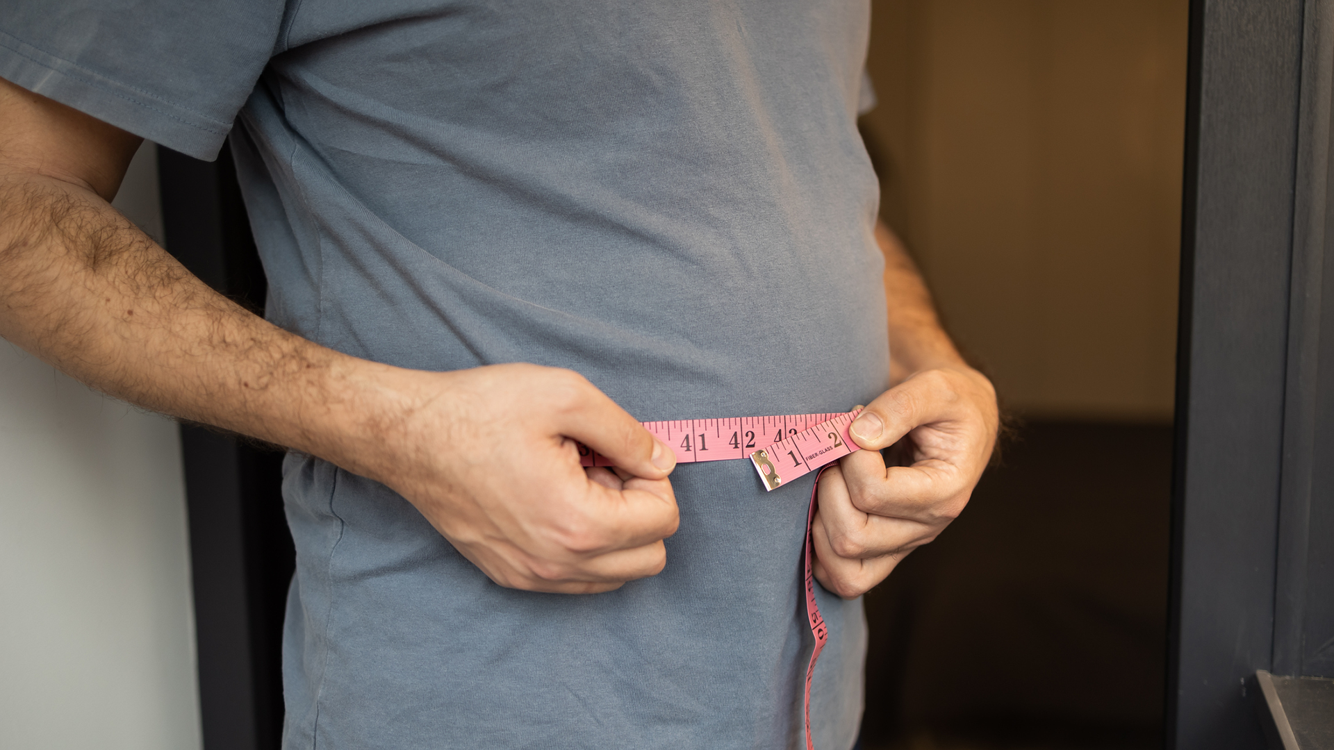 Close-up photograph of a person measuring their waist circumference with a pink tape measure. The tape reads approximately 41–42 inches at the point where the ends meet. The person is wearing a grey t-shirt and the shot is cropped to show only the torso an