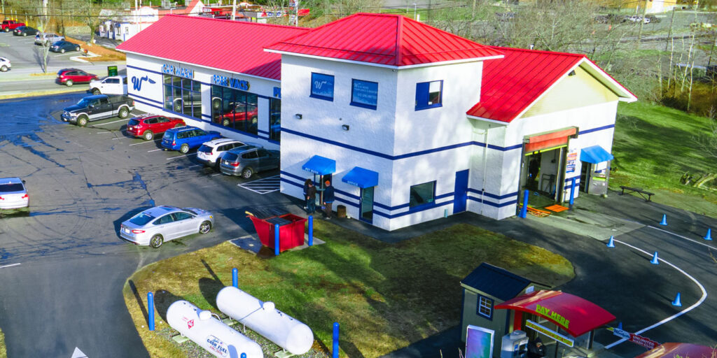 Car wash facility with red roof, multiple vehicles parked, and blue awnings, surrounded by roads and greenery.