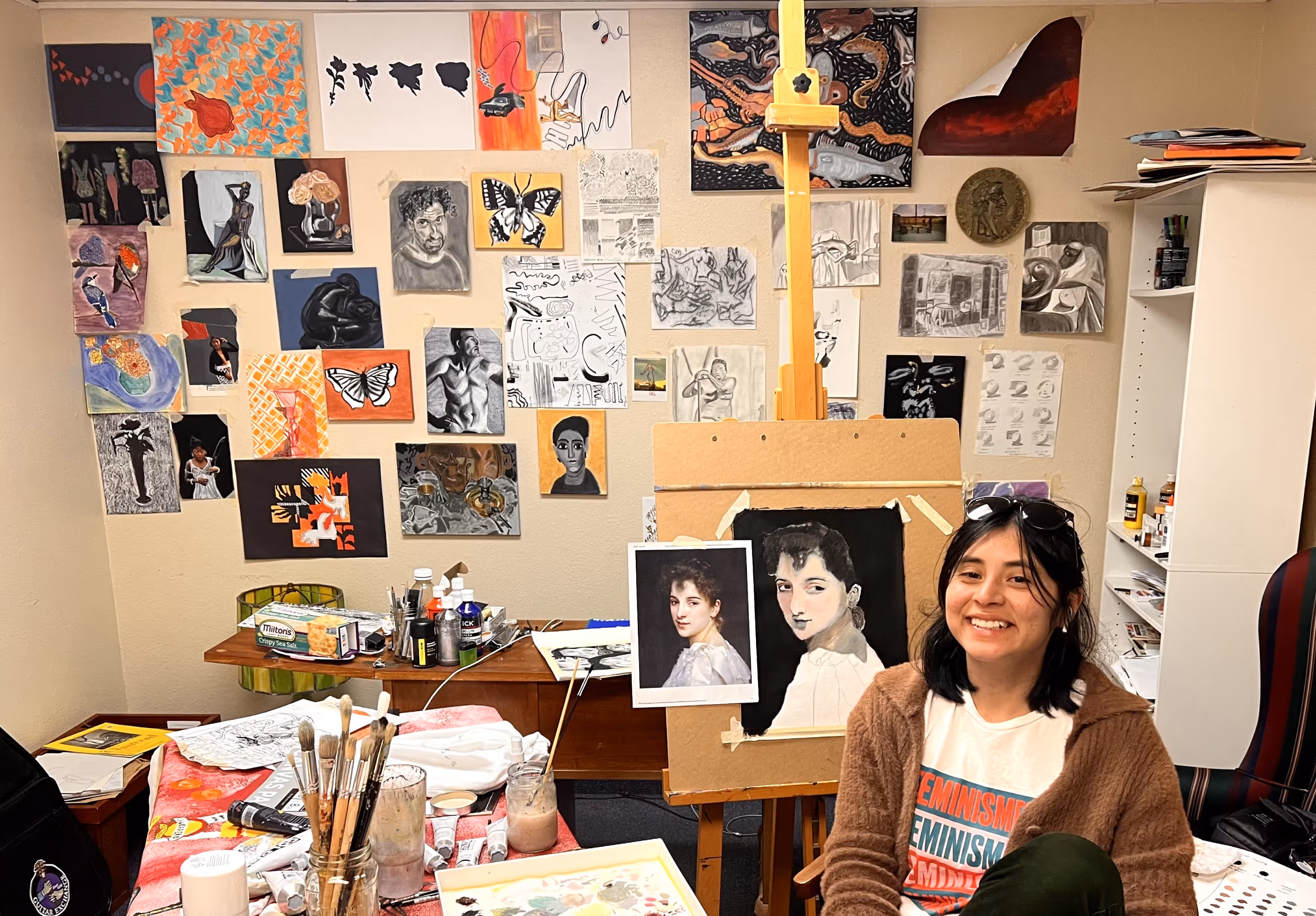 Claudia H., a portfolio program student, smiling in front of a portrait she painted in one of our courses. Behind her is a wall decorated with about 30 artworks she created in our classes.
