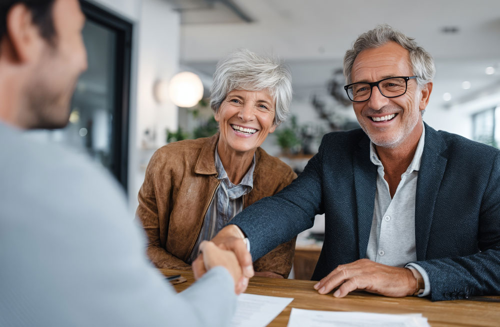 Smiling elderly couple shaking hands with a younger person across a table in a modern office.