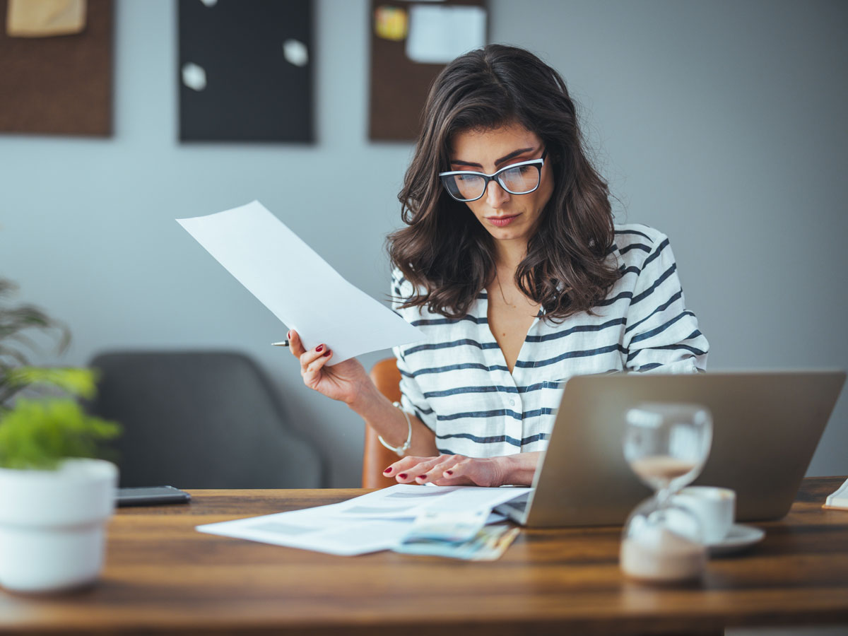 Woman with glasses and striped shirt reviewing documents at a wooden desk with a laptop and hourglass.