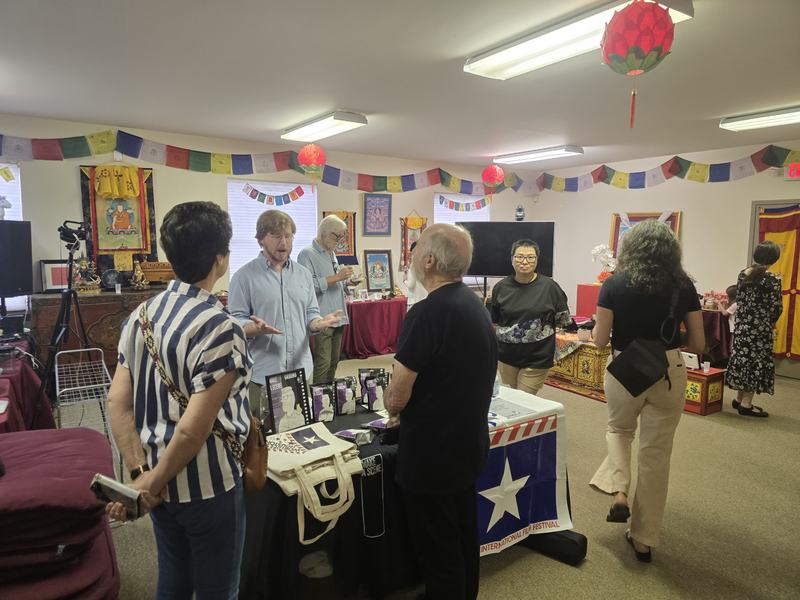 WorldFest-Houston merchandise table with tote bags and a festival banner, set up beneath Tibetan prayer flags at Dawn Mountain.