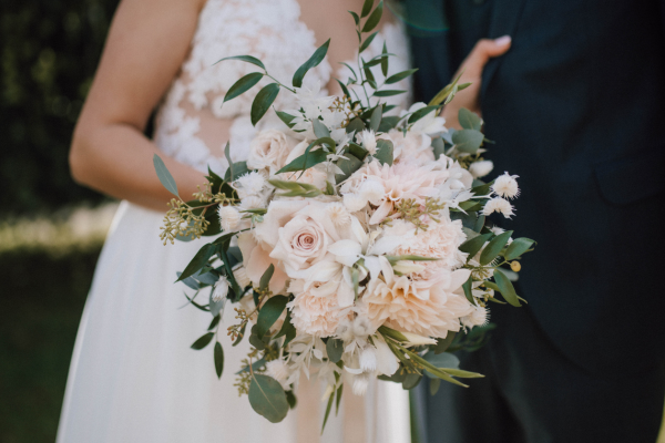 Bride holding a bouquet of cream roses, white flowers, and green foliage next to a groom in a dark suit.