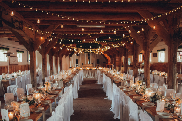 Rustic indoor reception with long tables, white-covered chairs, floral centerpieces, and string lights hanging from wooden beams.