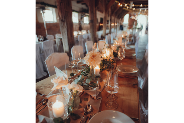Long wooden table set with white plates, glassware, candles, and floral centerpieces in a rustic indoor venue with wooden beams.