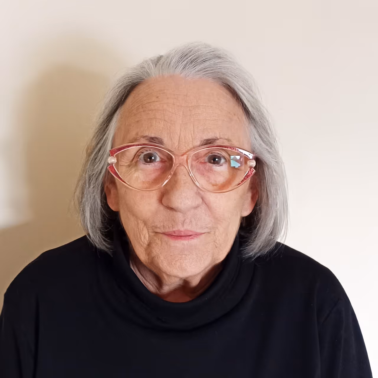 Portraitfoto von Frau Iris Schlüsener vor hellem Hintergrund, mit grauem Haar, roséfarbener Brille und schwarzem Rollkragenpullover.