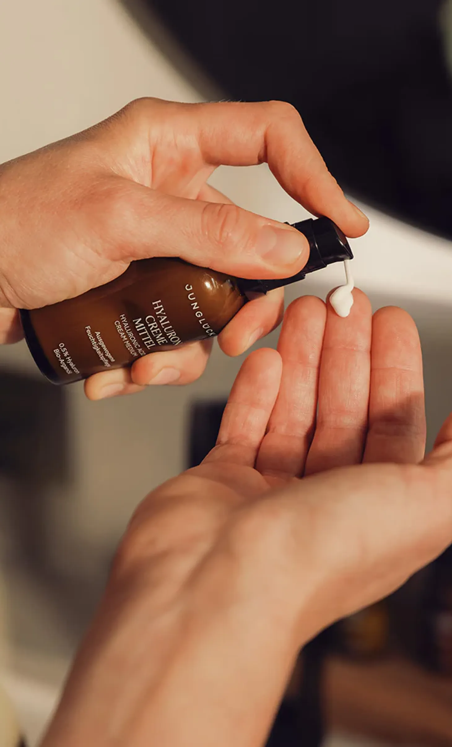 Hand dispensing white cream from a brown bottle labeled Junglück Hyaluron Creme Mittel onto a palm.