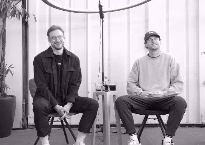 Two men sit facing each other in a studio with a microphone setup between them.