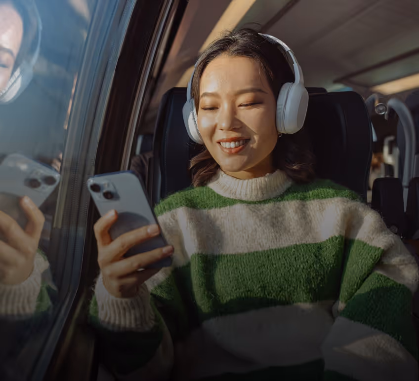 A woman wearing a green and white striped sweater and white headphones is sitting by a window in a train or bus, smiling as she looks at her smartphone.